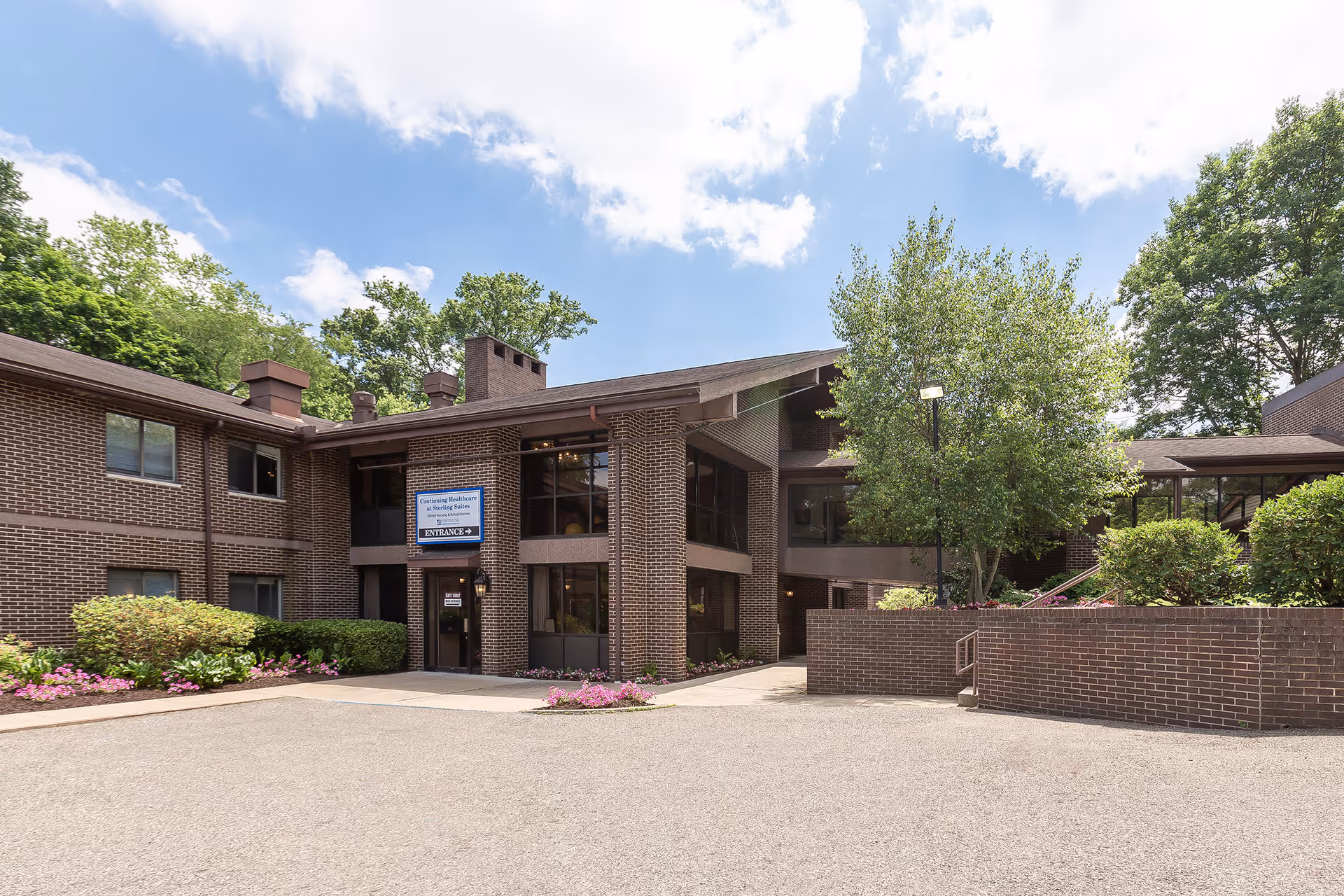 Exterior view of a two-story brick building with large windows, surrounded by greenery and flowering plants under a partly cloudy sky. A sign near the entrance reads 'Continuing Healthcare at Sterling Suites' with an arrow pointing to the entrance door.