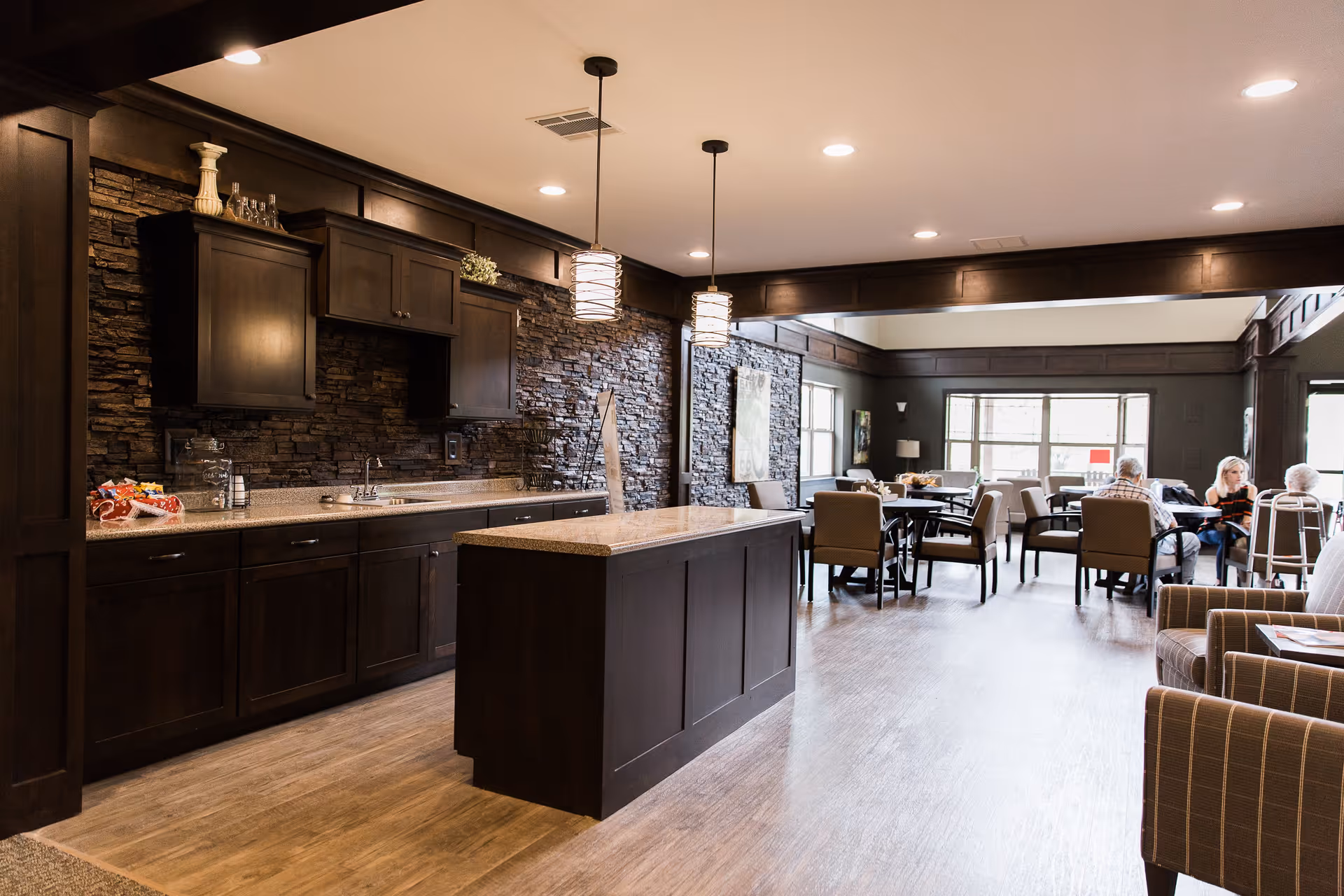 A spacious assisted living facility common area featuring a kitchen with dark wood cabinets, a stone backsplash, and a central island. The room extends into a dining area with several tables and chairs where elderly residents are seated and engaged in conversation. The space is well-lit with ceiling lights and pendant lamps, and has large windows allowing natural light to enter.