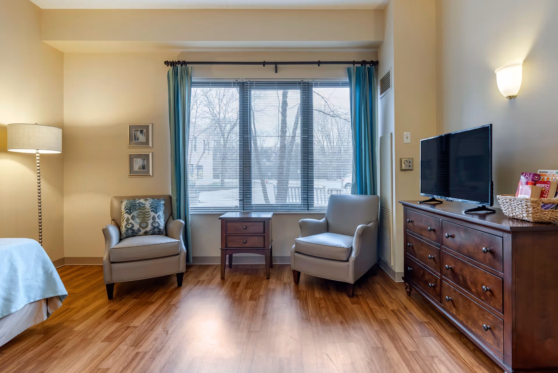 A cozy room with two gray armchairs separated by a small wooden side table in front of a large window with blue curtains and blinds. To the right, there is a wooden dresser with a flat-screen TV on top and a basket with magazines. On the left, part of a bed with a light blue bedspread and a tall floor lamp are visible. The room has wooden flooring and beige walls.
