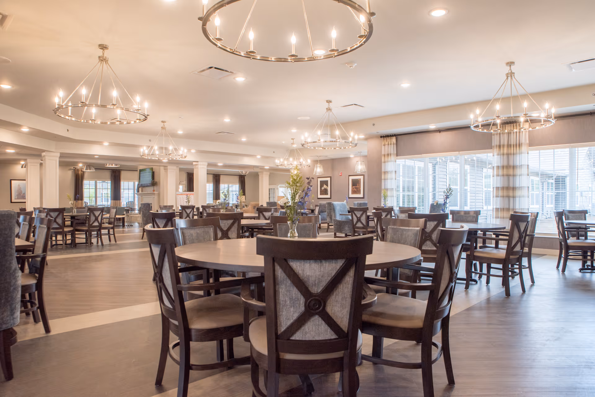 A spacious dining room in a senior living facility with multiple round and square wooden tables surrounded by cushioned chairs. The room is well-lit with large circular chandeliers hanging from the ceiling and natural light coming through large windows with striped curtains. There are decorative plants on some tables and framed artwork on the walls.