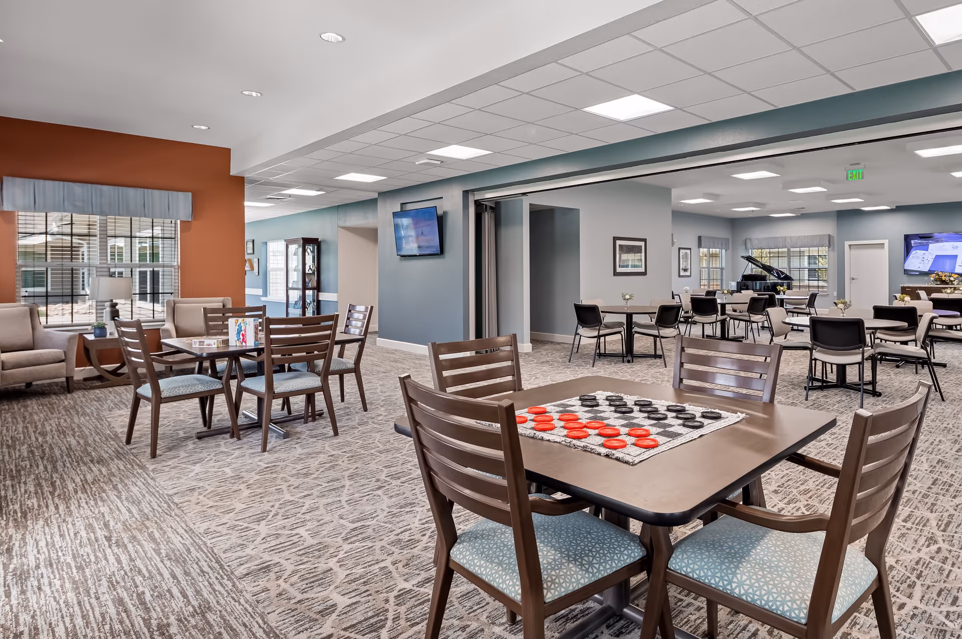 A spacious and well-lit common area in a senior living facility with multiple tables and chairs arranged for social activities. One table in the foreground has a checkers game set up. The room features carpeted floors, large windows with valances, a wall-mounted TV, and a grand piano in the background. The walls are painted in soft blue and orange tones.