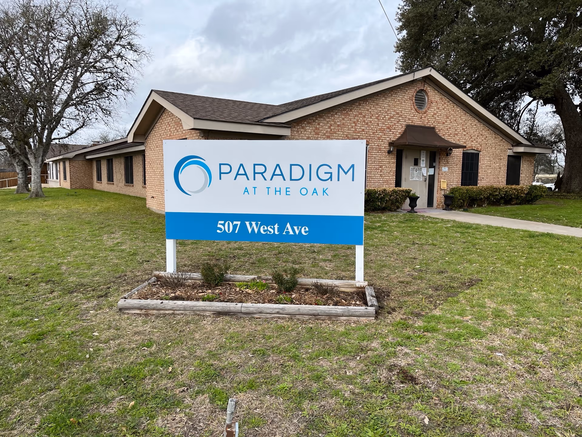 Exterior view of a brick building with a sign in front that reads 'Paradigm at the Oak, 507 West Ave'. The building has a small awning over the entrance door and is surrounded by grass and a few trees.