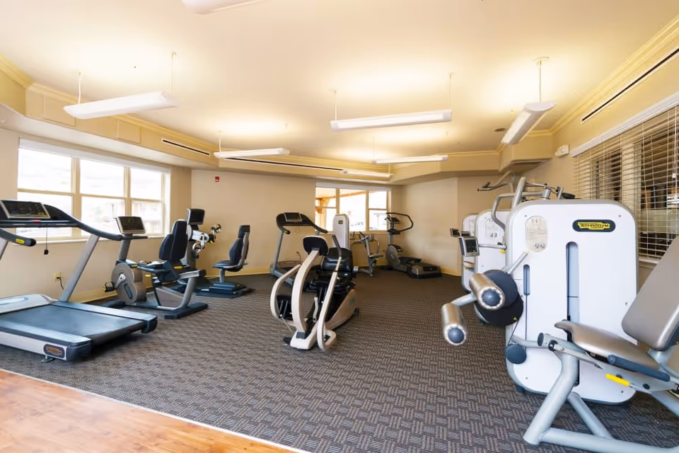A well-lit fitness room in a senior living facility featuring various exercise machines including treadmills, stationary bikes, and strength training equipment. The room has large windows allowing natural light to fill the space, beige walls, and patterned carpet flooring.