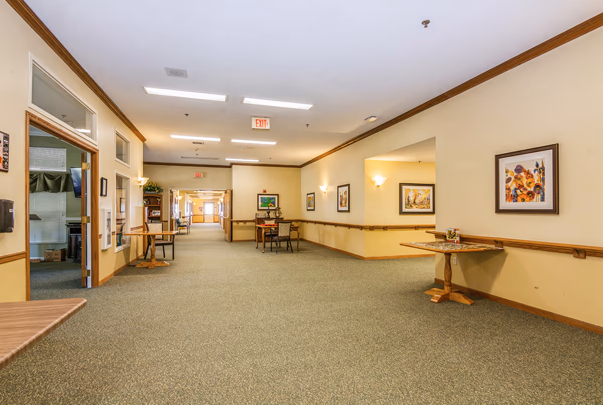 Wide hallway in a senior living facility with beige walls, green carpet, wooden trim, and several framed paintings on the walls. There are tables and chairs along the hallway, and an exit sign is visible on the ceiling. The hallway leads to a well-lit area in the distance.