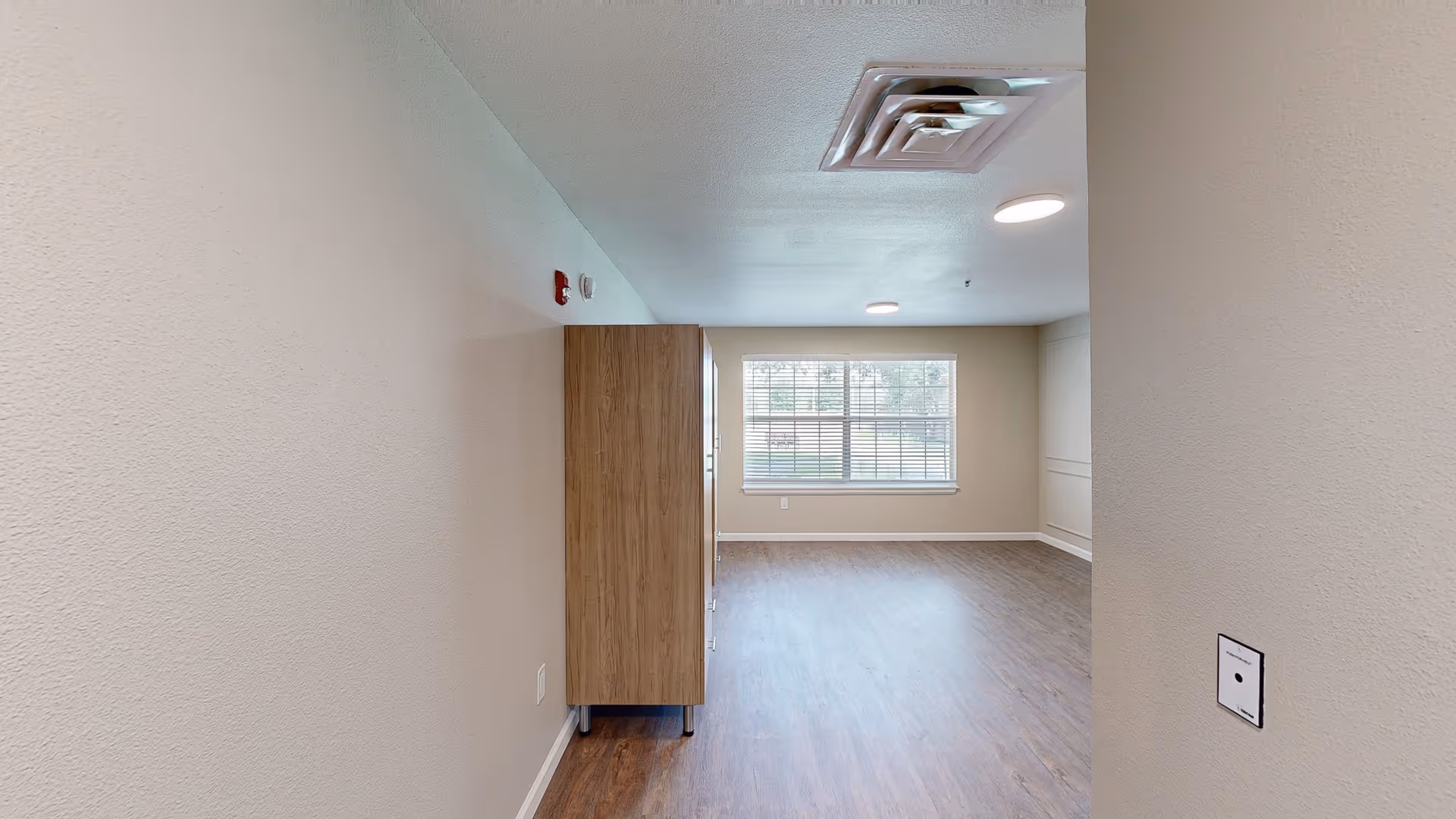 Empty room with wood flooring, beige walls, a large window with blinds, and a tall wooden cabinet against the left wall. The ceiling has a vent and round recessed lights.
