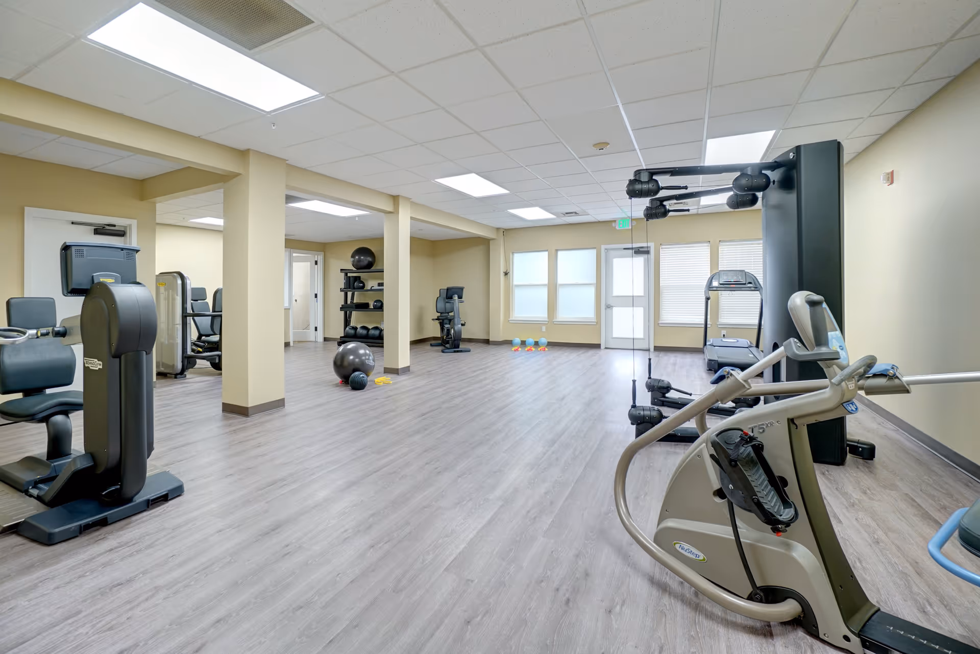 A spacious fitness room in a senior living facility with various exercise equipment including stationary bikes, a treadmill, weight machines, exercise balls, and dumbbells. The room has light-colored walls, large windows with blinds, and a light wood floor.