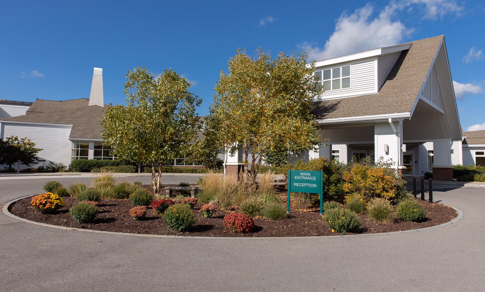 Exterior view of a senior living facility building with a covered entrance and a circular driveway. There is a landscaped roundabout with trees, shrubs, and flowers in front of the entrance. A green sign reads 'Main Entrance Reception'. The sky is clear and blue.