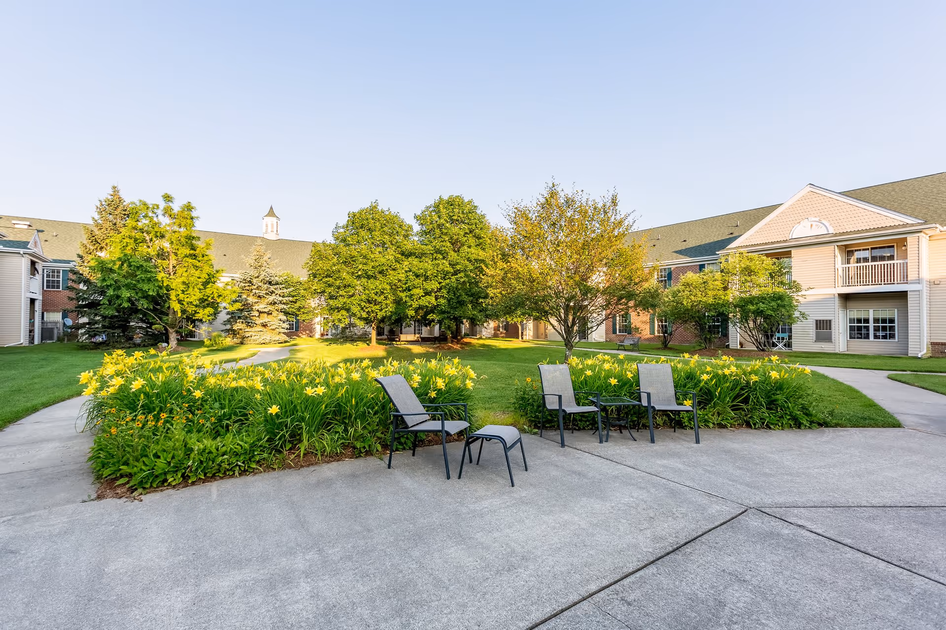 Outdoor courtyard area at Independence Village of Grand Ledge featuring a concrete patio with four chairs and a small table, surrounded by green grass, yellow flowers, and trees, with two-story residential buildings in the background under a clear sky.