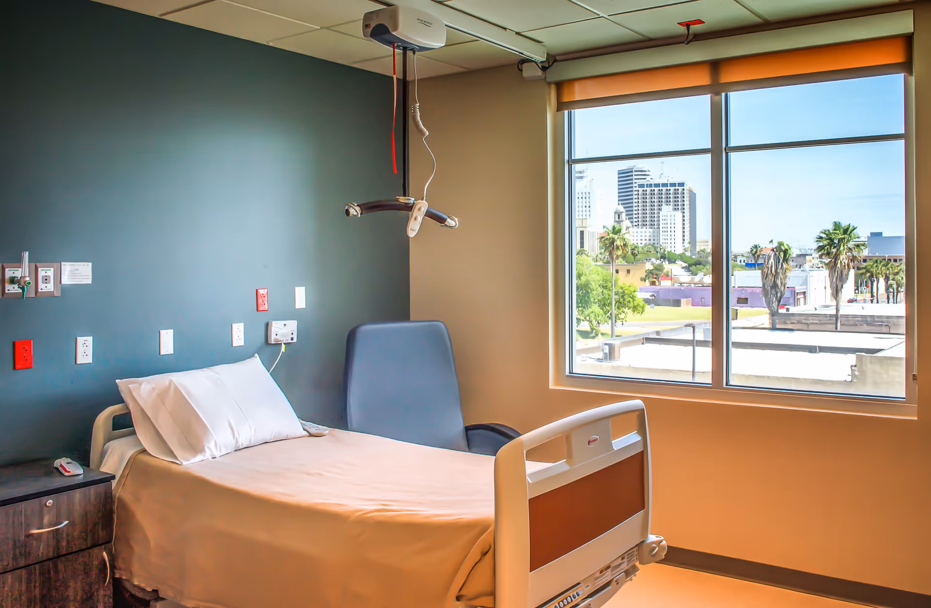 A hospital room with a single bed made with white sheets and a pillow, a bedside table with a call button, a blue chair, and a large window showing a cityscape with palm trees and buildings outside.