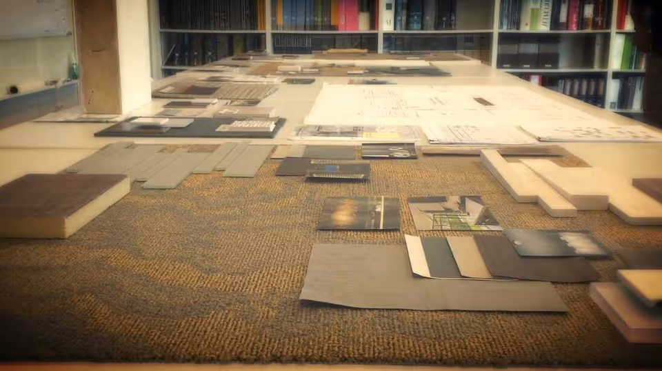 A close-up view of various architectural materials, samples, and blueprints laid out on a table, with shelves of books in the background.