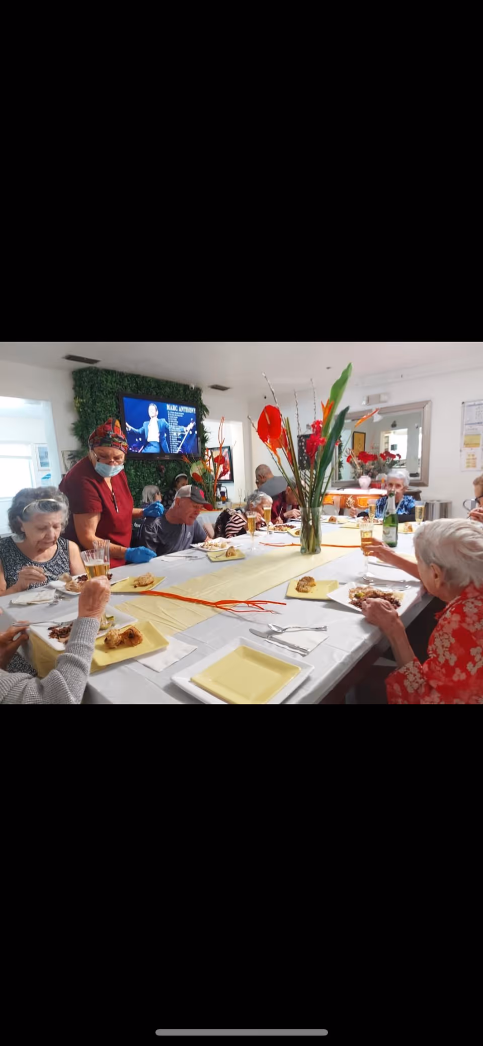 Elderly residents seated around a long communal dining table sharing a meal with a staff member serving and a tall floral centerpiece.