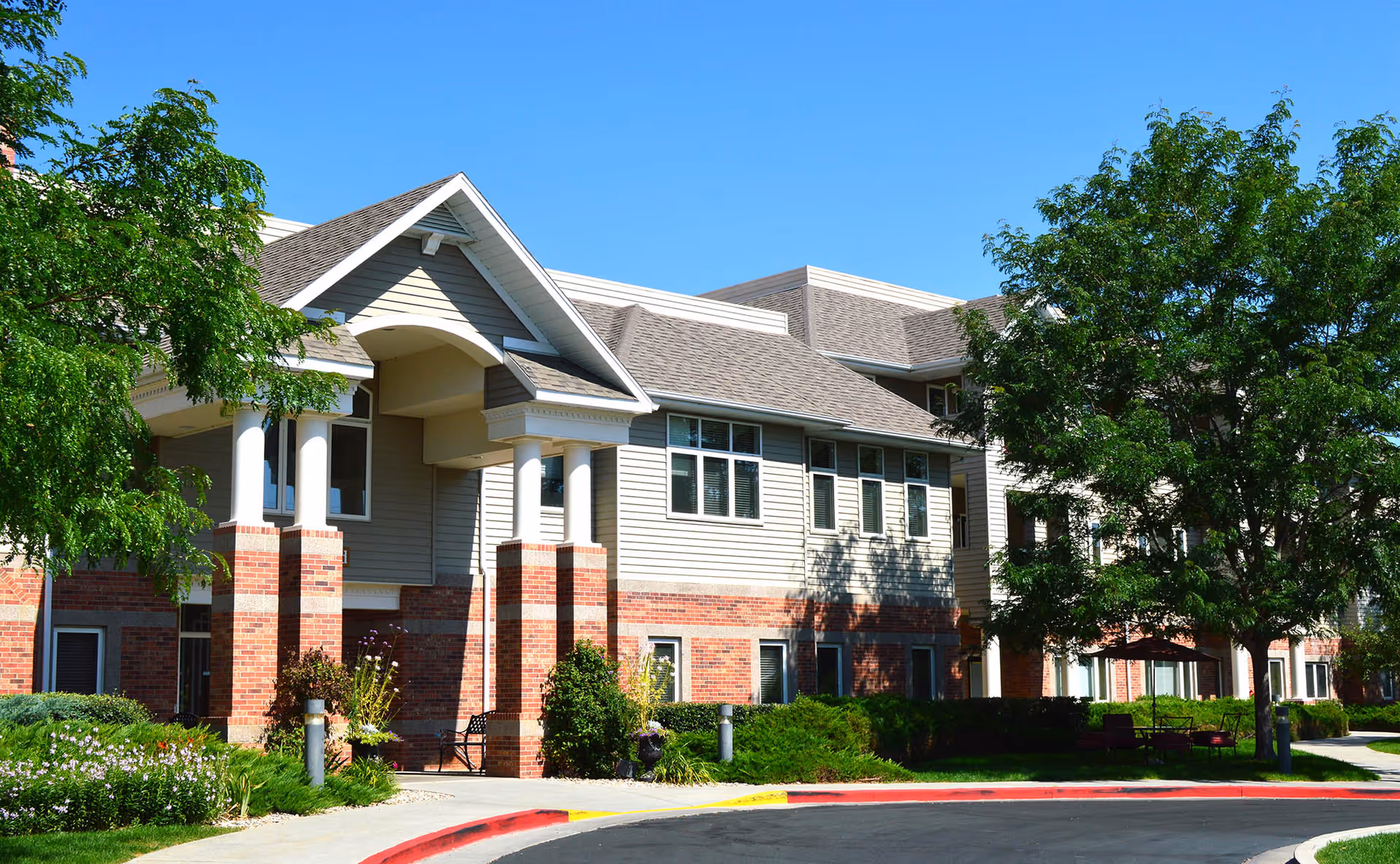 Exterior view of a senior living facility named The Wexford, showing a two-story building with a combination of brick and siding, large windows, a covered entrance with columns, surrounded by green trees and landscaping under a clear blue sky.