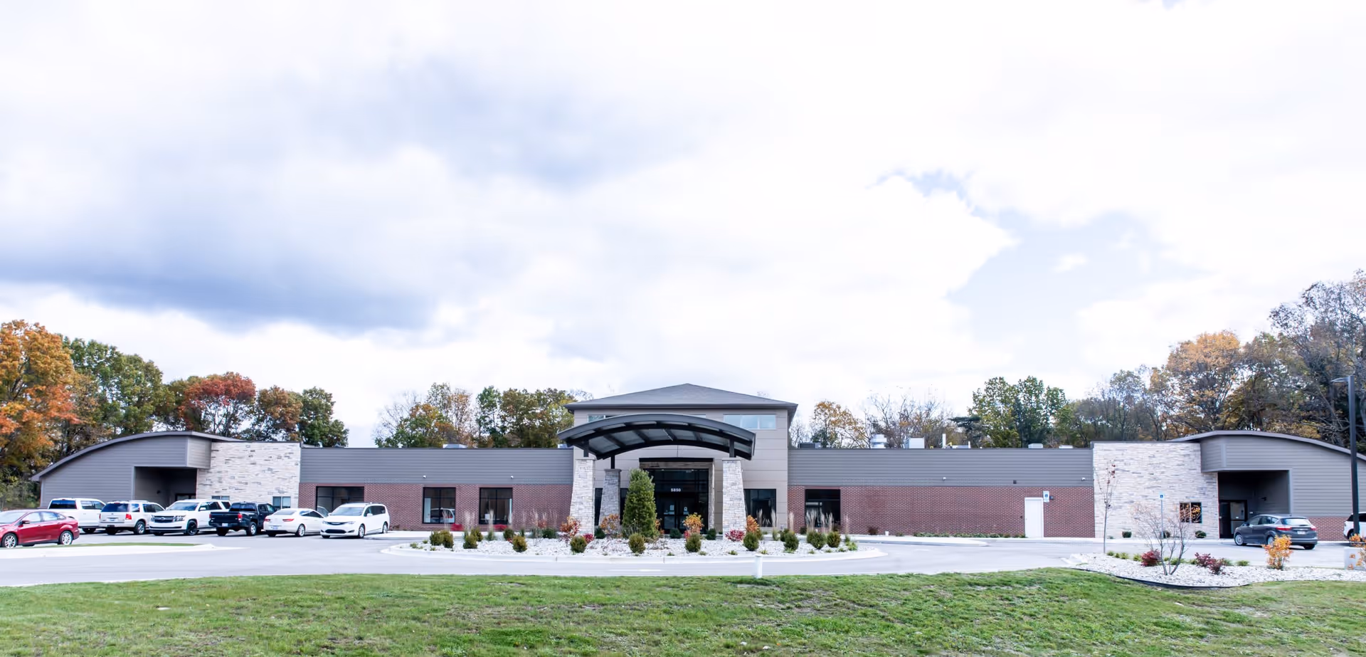 Front exterior view of a single-story senior living facility building with a covered entrance, surrounded by a parking lot with several parked cars and trees with autumn foliage in the background under a partly cloudy sky.