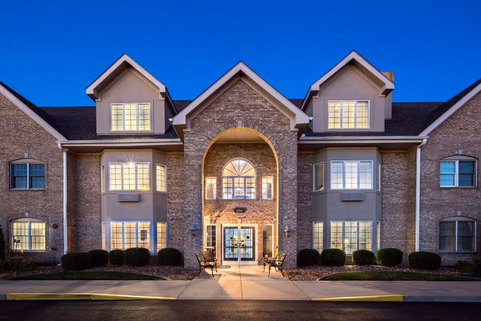 Front entrance of a two-story brick senior living building lit at dusk with benches and trimmed shrubs.