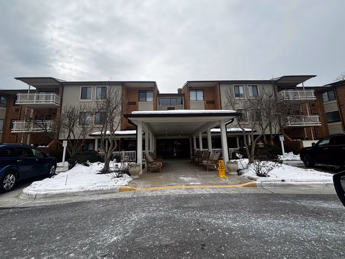 Front entrance of a three-story assisted living building with a covered driveway, benches and snow on the ground.