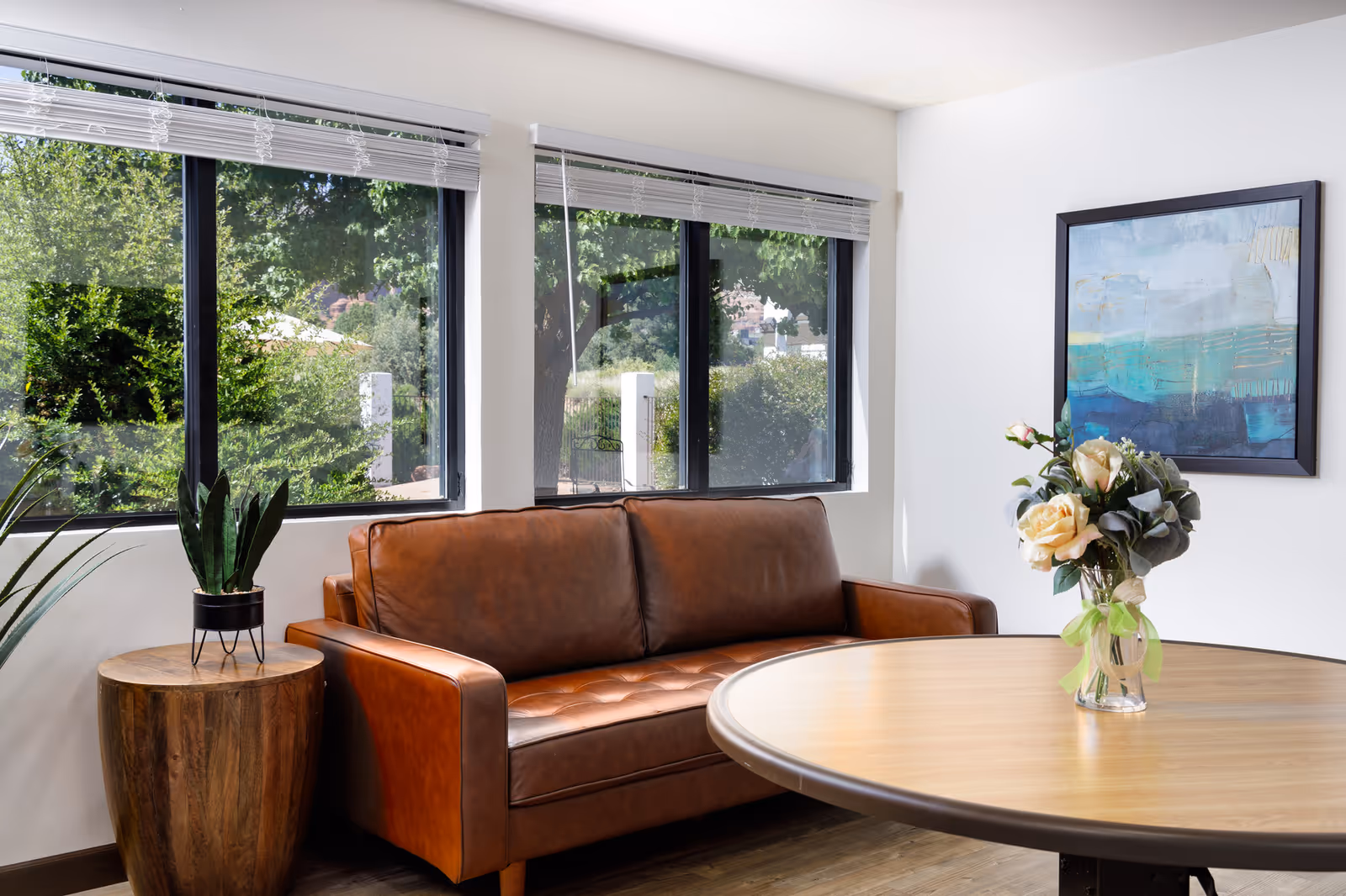 A cozy interior room with a brown leather sofa, a round wooden table with a vase of flowers, a wooden side table with a potted plant, and large windows showing green trees outside. A framed abstract painting hangs on the white wall.