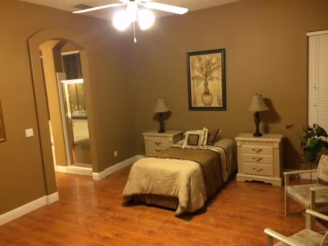 A cozy bedroom with a single bed covered in beige and brown bedding, flanked by two matching nightstands each with a lamp. A framed artwork of a tree hangs on the wall above the bed. The room has wooden flooring, beige walls, a ceiling fan with lights, and an open doorway leading to a bathroom area with a glass shower enclosure. There is also a chair with a cushion and a potted plant near the window with closed blinds.