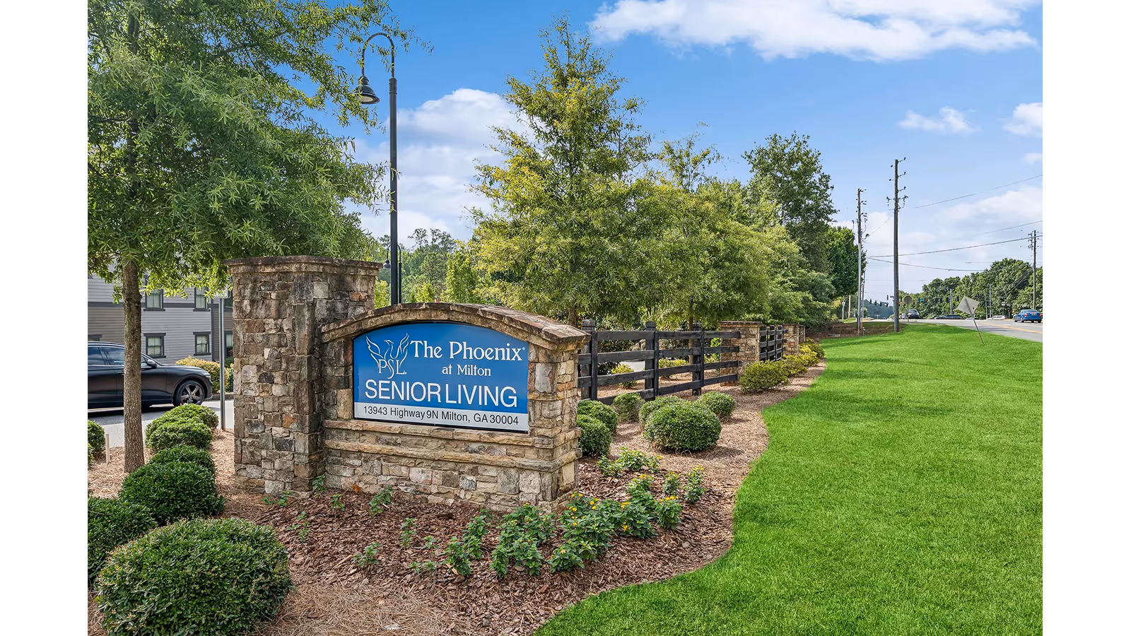 Stone entrance sign for The Phoenix at Milton senior living surrounded by landscaping and a green lawn by the roadside.