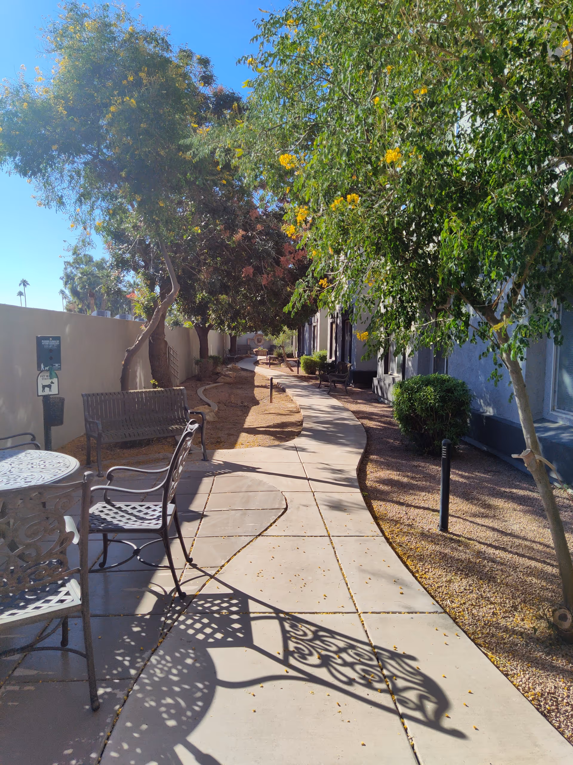 A sunny outdoor walkway at Ridge at the Stratford with metal chairs and tables on the left side, benches along the path, and trees providing shade. The path curves gently and is bordered by gravel and landscaping.