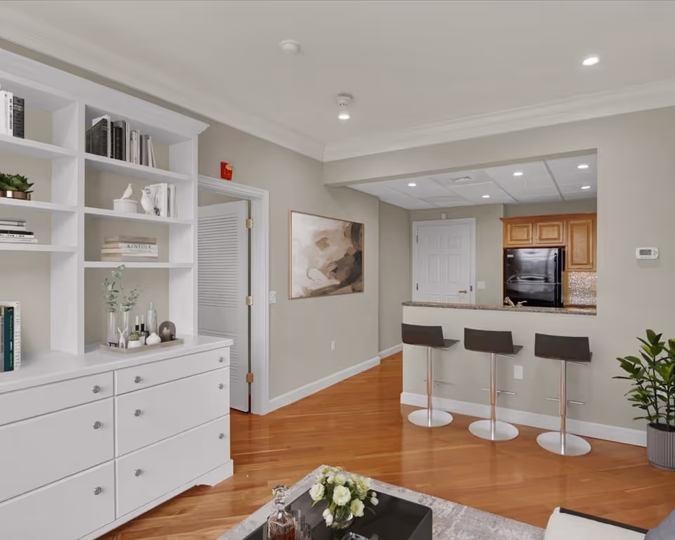 Interior view of a senior living community featuring a white built-in bookshelf with books and decorative items on the left, a kitchen area with wooden cabinets and a black refrigerator in the background, and a breakfast bar with three modern bar stools. The room has wooden flooring, neutral-colored walls, a piece of abstract art on the wall, and a small plant in a pot on the right side.