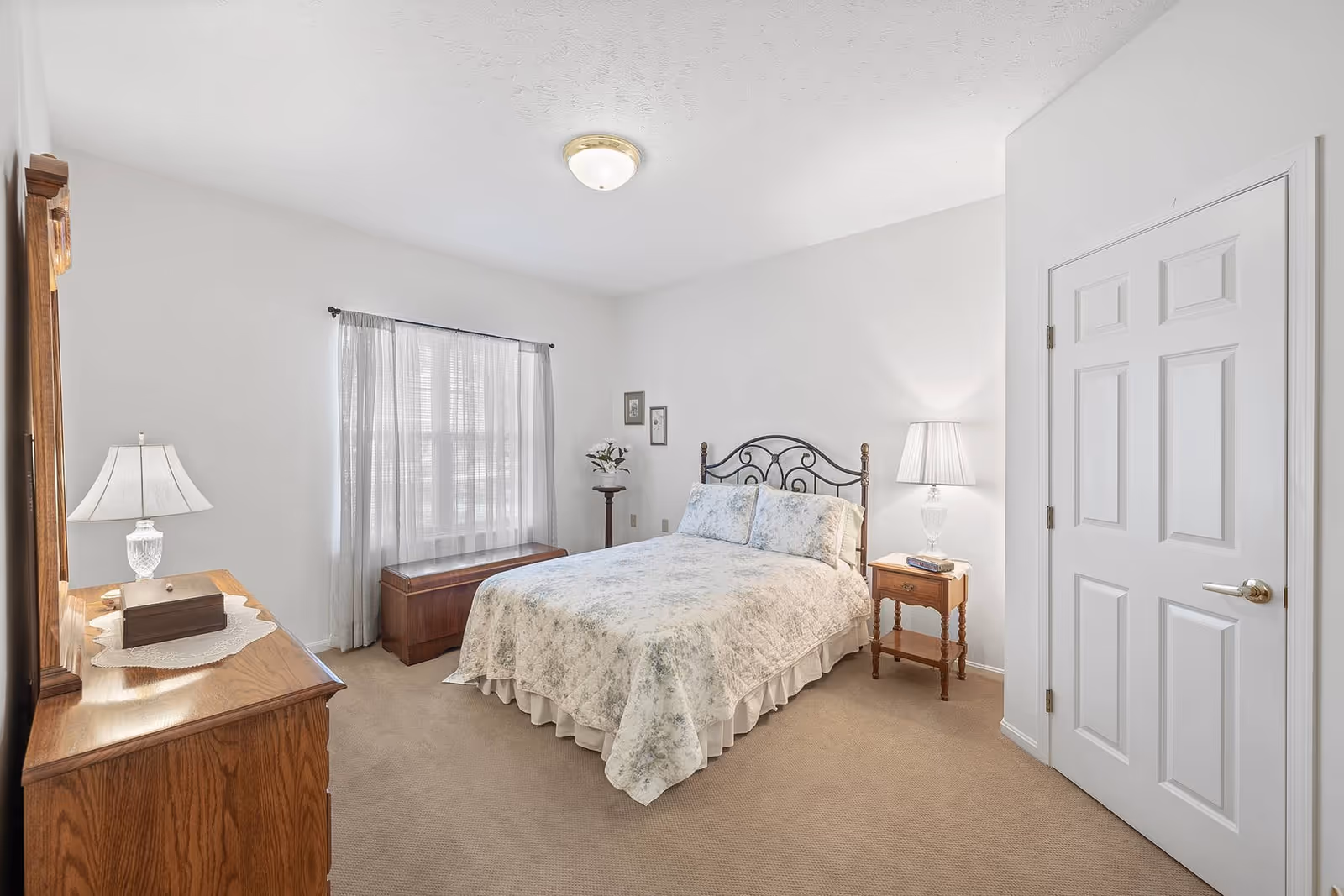 A bright bedroom with a double bed covered in a floral quilt, a wooden dresser with a lamp and decorative box, a wooden chest under a window with sheer curtains, a small wooden nightstand with a lamp and a book, and a closed white door.