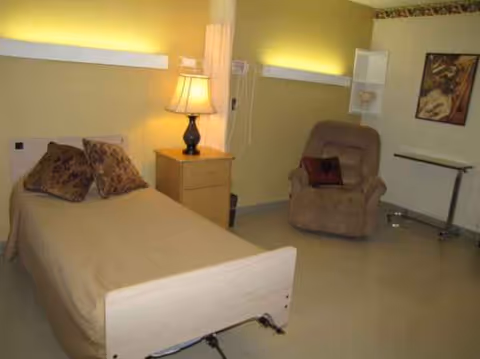A small bedroom with a single bed, nightstand, lamp, and beige recliner chair.