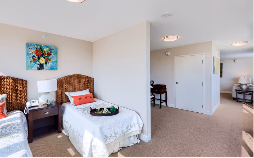 A bright room with two twin beds featuring wicker headboards and white bedspreads with floral patterns. A nightstand with a lamp, telephone, and small clock is placed between the beds. A colorful floral painting hangs on the wall above one bed. The room opens into a hallway with a closed door, a small desk with a chair, and a living area with a sofa and side table with a lamp visible in the background.