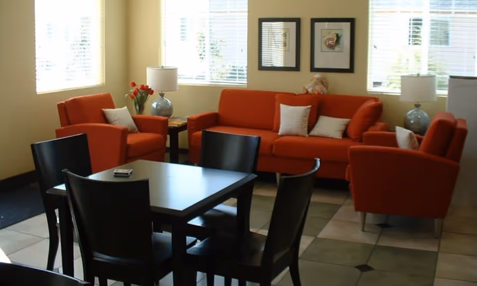 A cozy living room area with an orange sofa and two matching armchairs, each with white cushions. There are two blue table lamps on side tables next to the sofa, a vase with red flowers, and two framed pictures on the wall. In the foreground, there is a dark wooden dining table with four chairs. The room has large windows with white blinds letting in natural light.