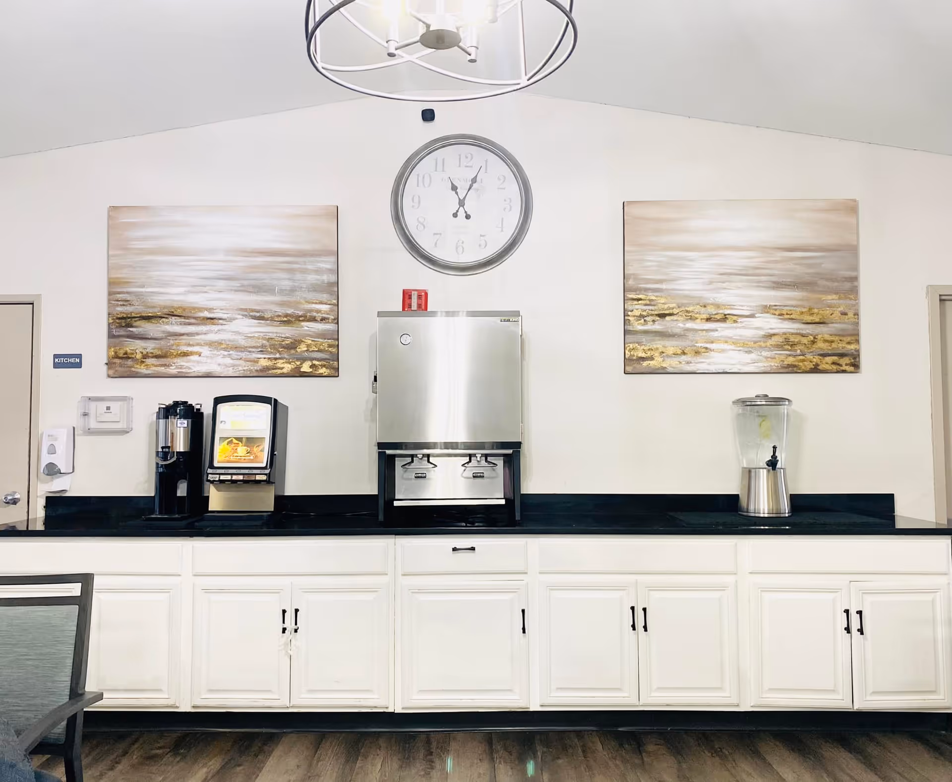 A beverage station in a room with white cabinets and a black countertop. The station includes a coffee dispenser, a hot water dispenser, and a glass container with a spout filled with water and lemon slices. Above the station are two abstract paintings with gold and beige tones and a large round clock showing the time as 11:55. The walls are white, and there is a modern circular light fixture hanging from the ceiling.