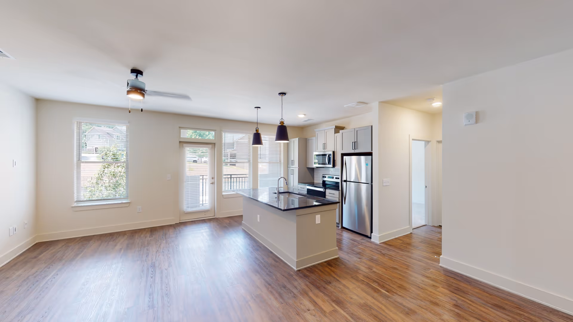 Bright and modern open-concept kitchen and living area with wood flooring, white walls, a ceiling fan, large windows, a glass door leading outside, and a kitchen island with a black countertop and two pendant lights above. Stainless steel appliances including a refrigerator, microwave, and stove are visible.