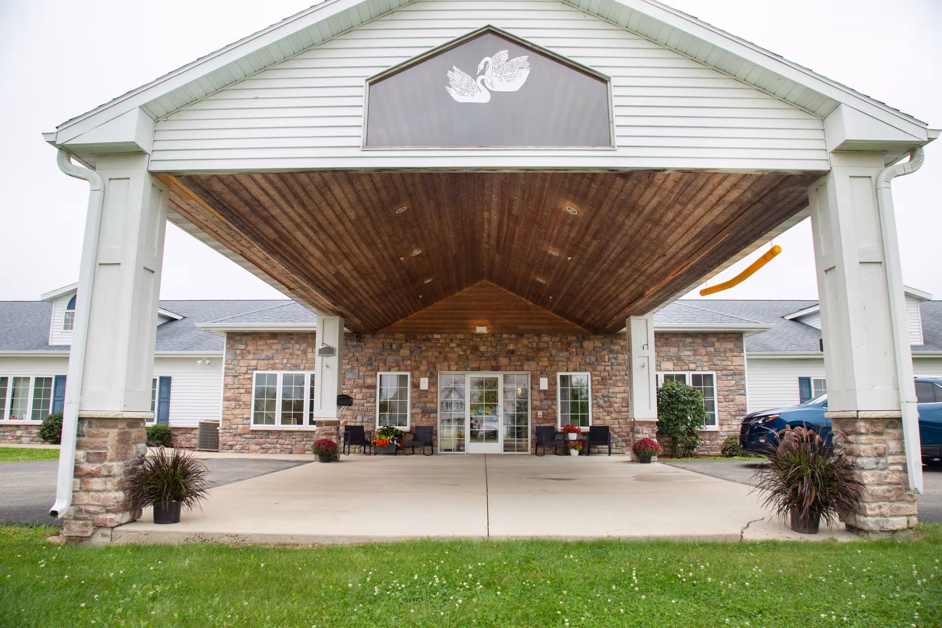 Entrance of Charleston House Assisted Living featuring a covered driveway with a wooden ceiling, stone facade, glass double doors, potted plants, and outdoor seating.