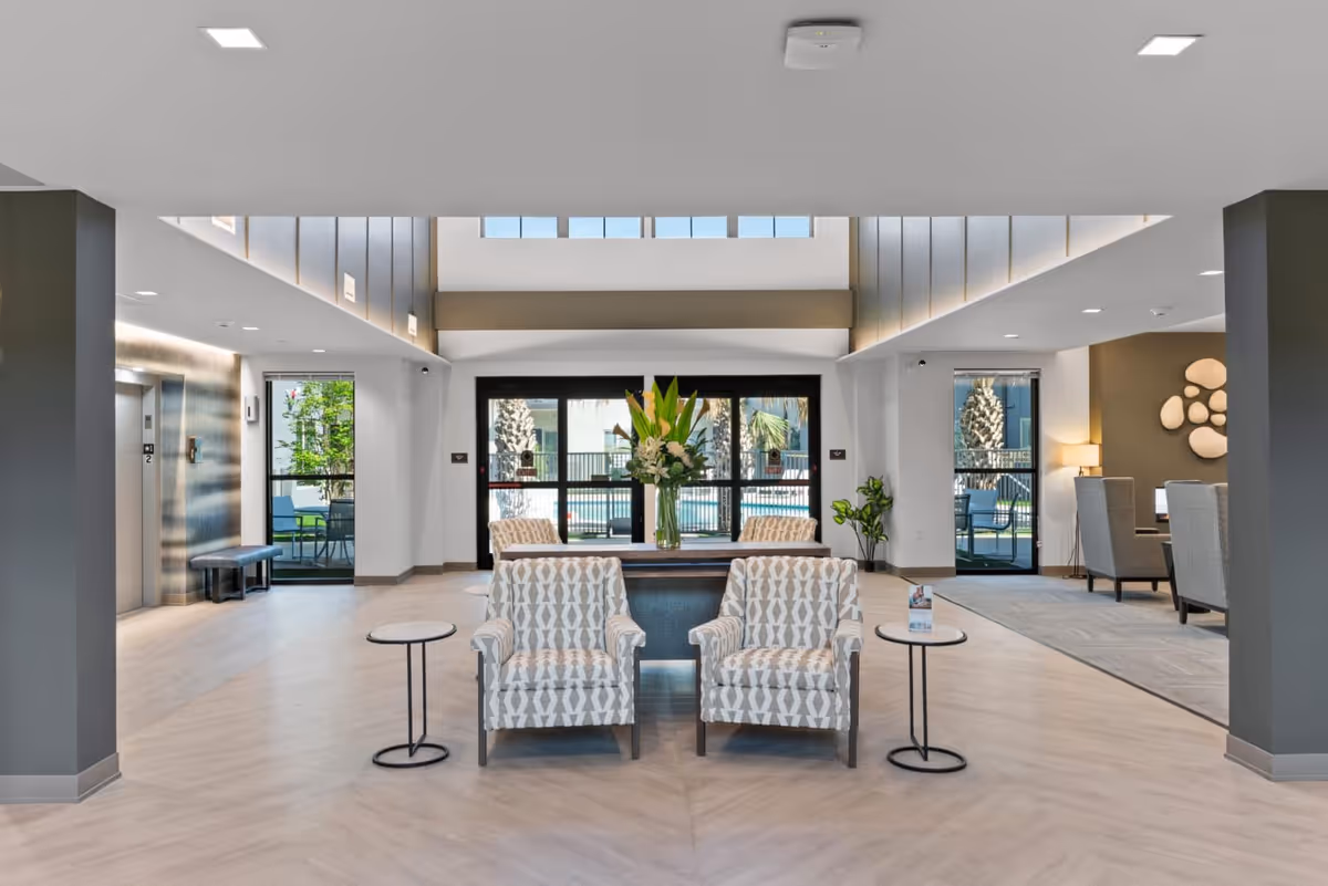 A spacious and modern lobby area with patterned armchairs arranged around a central table holding a large vase of flowers. The room features light wood flooring, large windows and glass doors leading outside, and a high ceiling with skylights. There are small round side tables next to the chairs, an elevator on the left, and a seating area with armchairs and a lamp on the right.