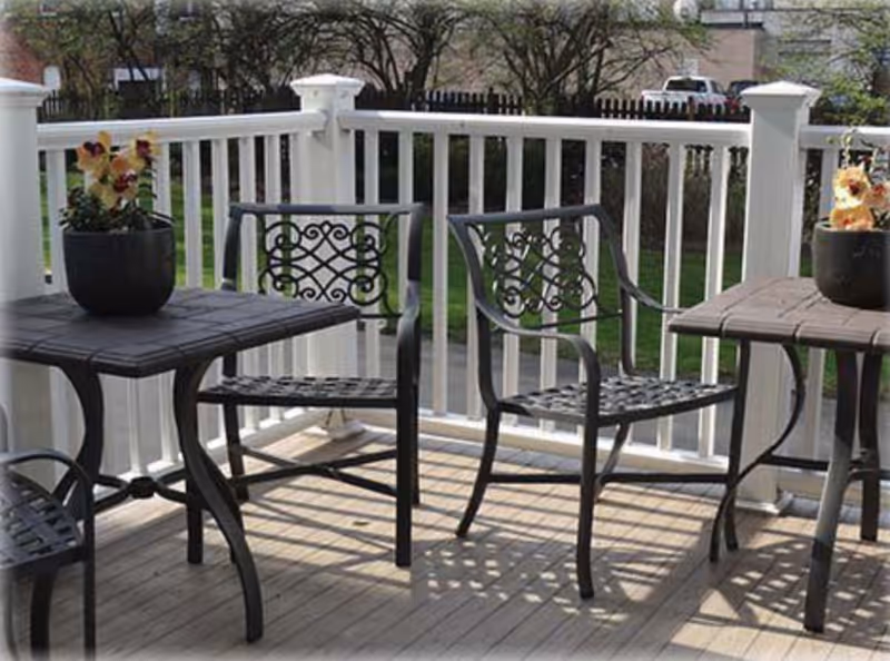 Outdoor patio area with two black metal chairs and two matching tables, each table holding a potted plant with flowers. The patio has a white railing and overlooks a green lawn with trees and parked vehicles in the background.
