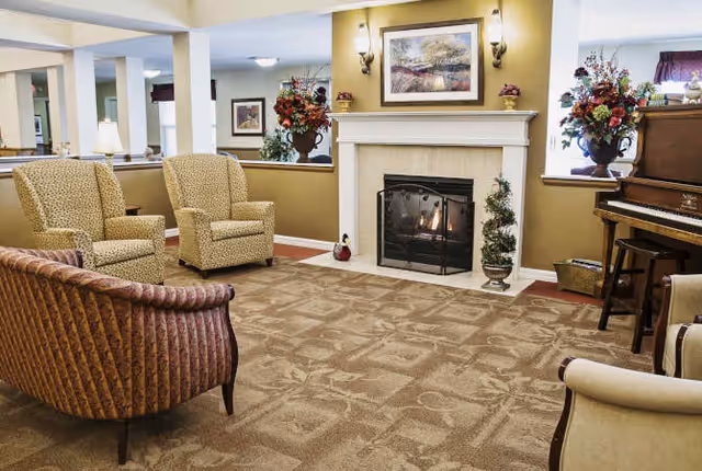 A cozy living room area with a lit fireplace in the center, flanked by two wall sconces and a framed landscape painting above the mantel. The room features patterned carpet, two beige armchairs, a brown patterned sofa, a piano with a stool, and floral arrangements on either side of the fireplace.