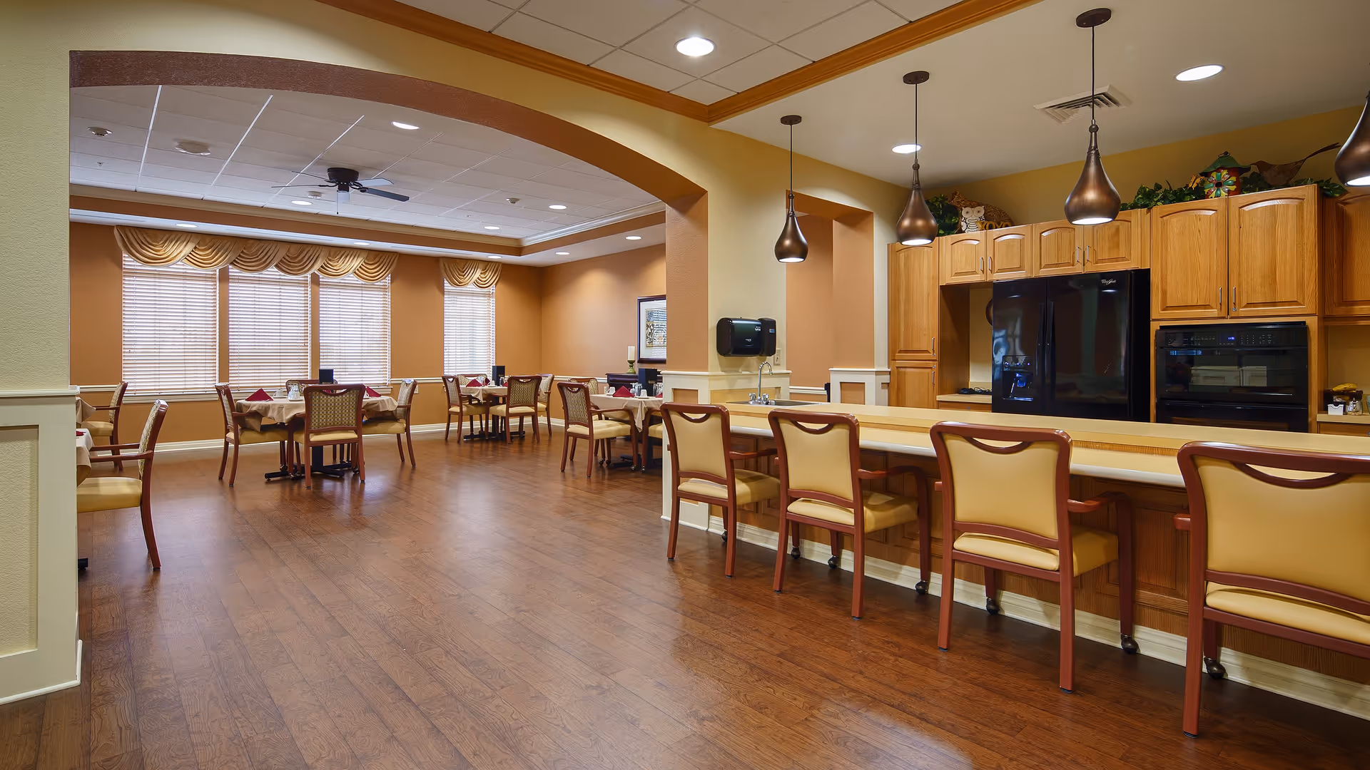 A spacious dining area in a senior living facility with wooden floors, several tables with chairs, and a kitchen counter with high chairs. The kitchen area has wooden cabinets, a black refrigerator, and pendant lights hanging from the ceiling. Large windows with blinds and valances allow natural light into the room.