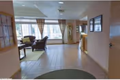 Interior view of a senior living facility common area with tiled floors, a seating area with armchairs and a coffee table near large windows with curtains, a grandfather clock, and framed pictures on the walls.