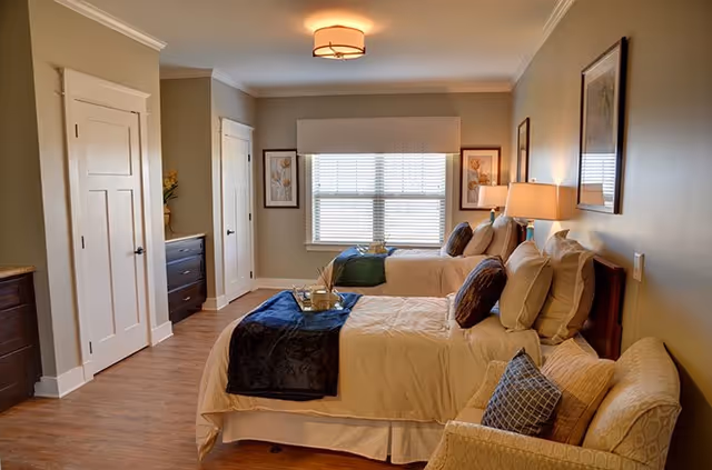 A cozy senior living bedroom with two twin beds neatly made with beige bedding and dark blue throws. The room features wooden flooring, a window with blinds, framed artwork on the walls, a cushioned armchair with pillows, and bedside lamps providing warm lighting. There are two white doors and a dark wooden dresser along one wall.