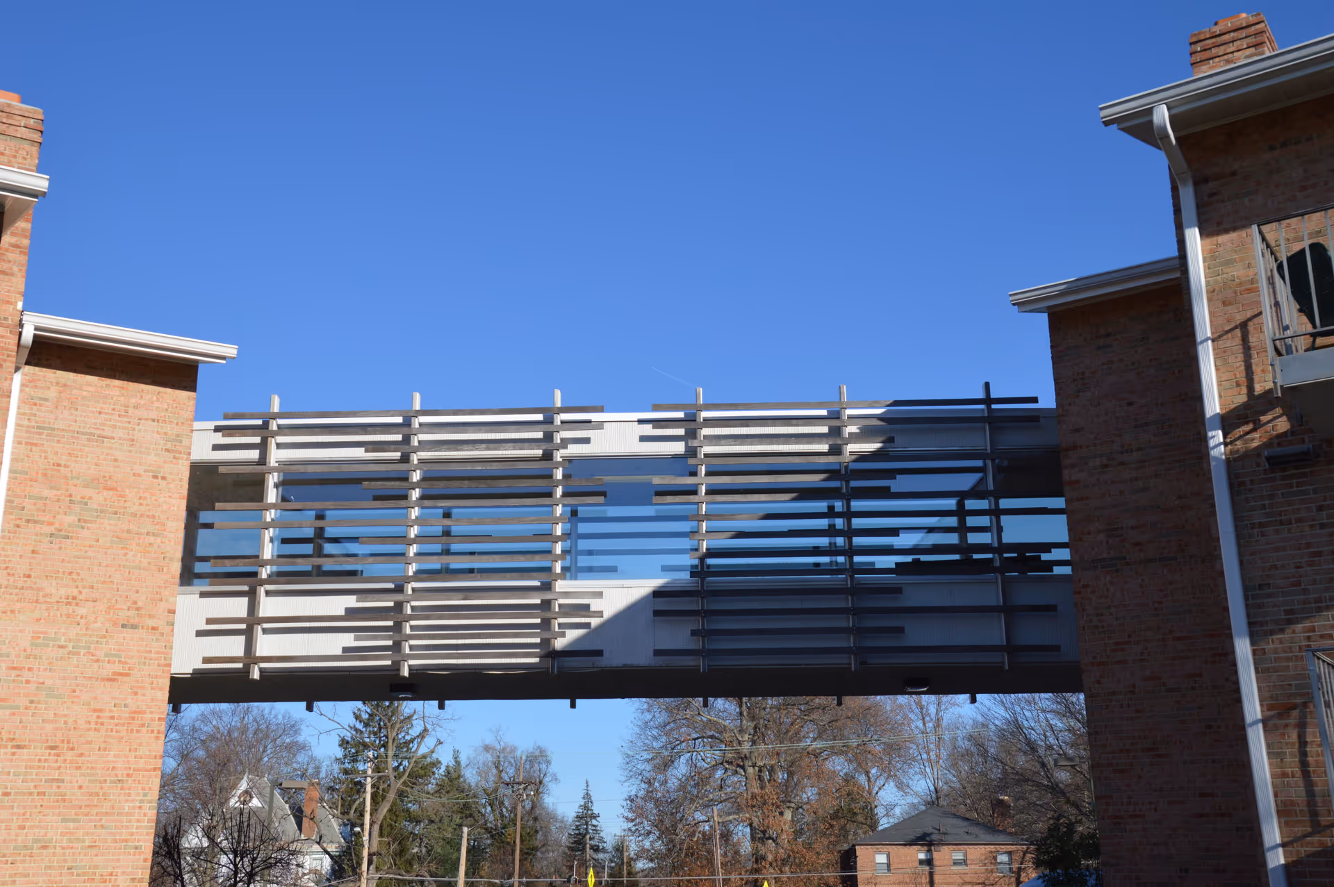 A glass-enclosed skybridge with horizontal sunshades connecting two brick buildings against a clear blue sky.