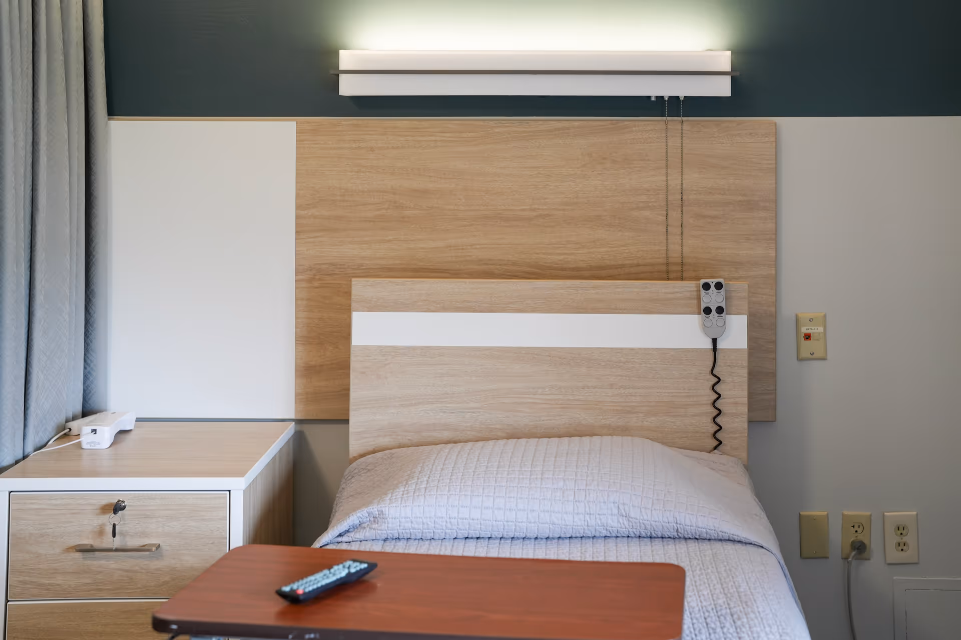 A single bed with a light wood headboard and a white and light blue quilted blanket in a senior living facility room. Next to the bed is a matching light wood nightstand with a white phone on it. Above the headboard is a horizontal light fixture. A remote control is placed on a brown overbed table in front of the bed. The wall behind the bed is painted in two tones, with a darker upper section and a lighter lower section. There are electrical outlets and a call button on the wall to the right of the bed.
