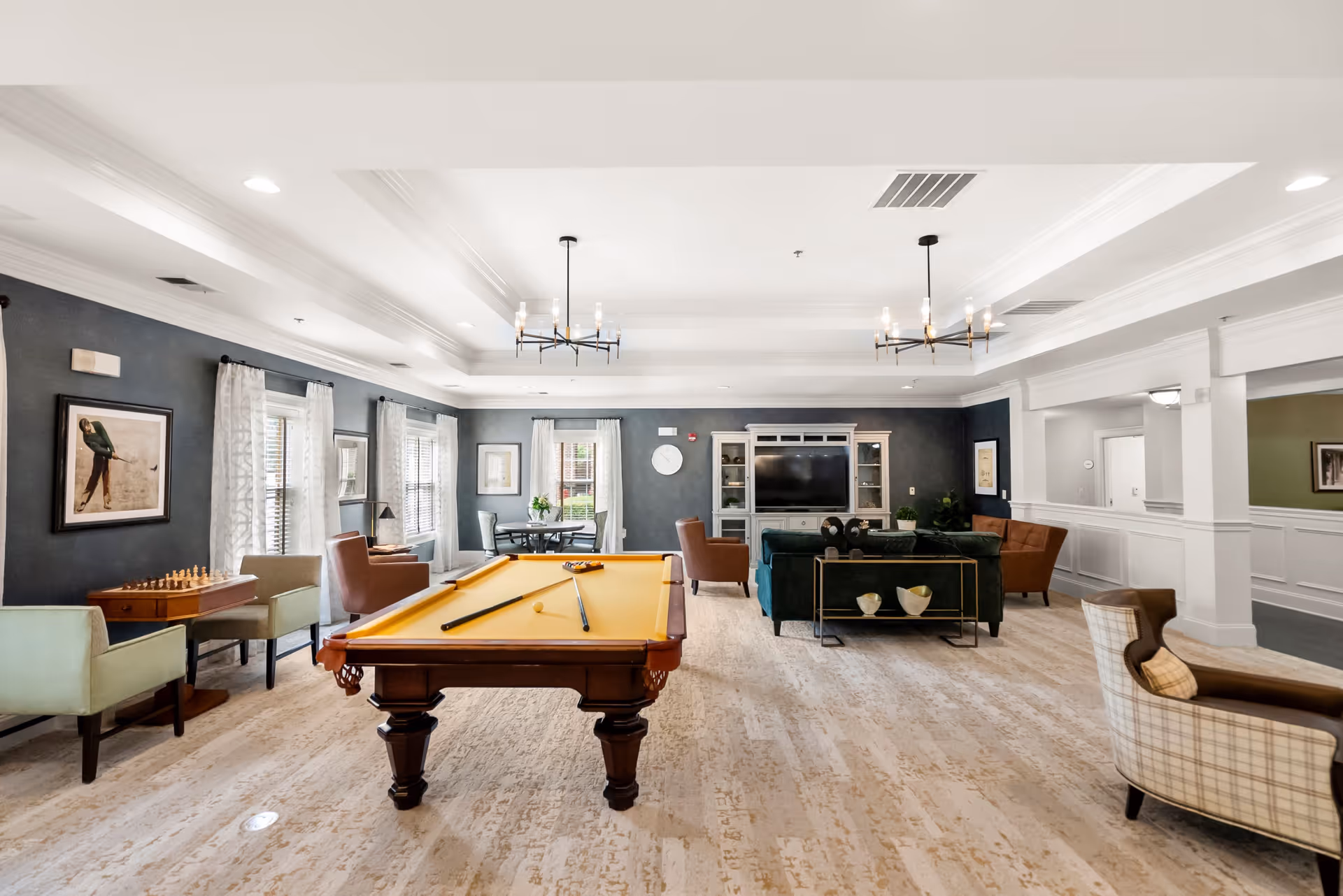 A spacious and well-lit common room featuring a pool table with cues, several armchairs, a chess table, and a seating area with a sofa and chairs facing a large TV. The room has light-colored flooring, dark accent walls with framed artwork, and large windows with white curtains allowing natural light to enter. Two modern chandeliers hang from the ceiling.