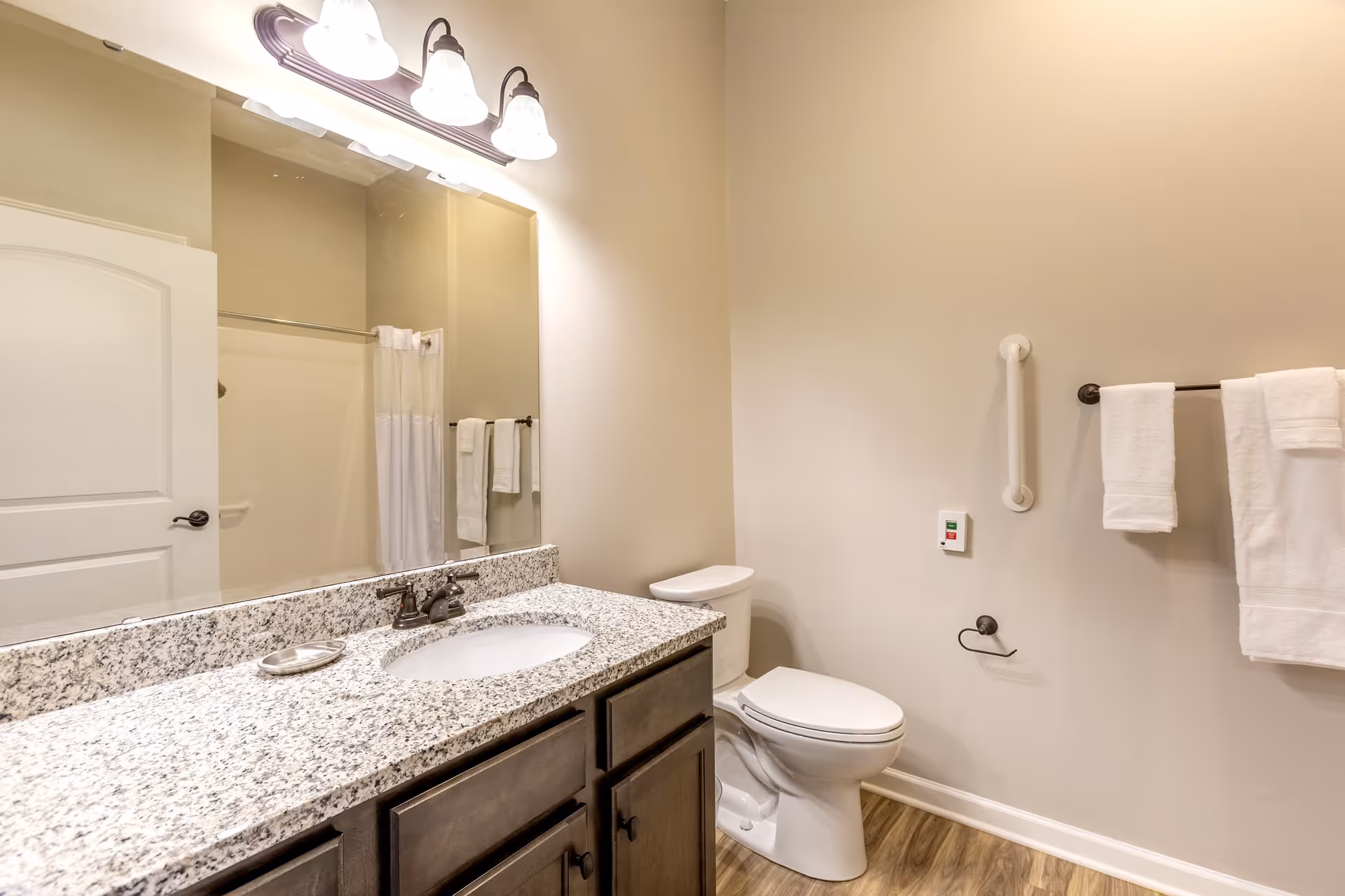 A clean and modern bathroom with a granite countertop sink, a large mirror, a toilet, and a towel rack with white towels. The walls are painted beige and the floor has wood-style flooring. There is a white door partially visible on the left side.