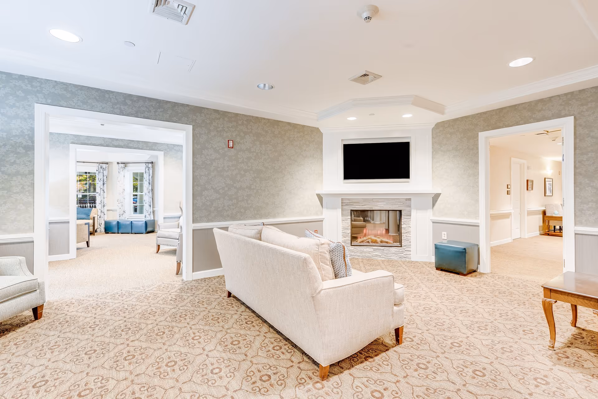 Bright lounge area with sofas facing a fireplace and wall-mounted TV, with doorways leading to additional seating rooms.