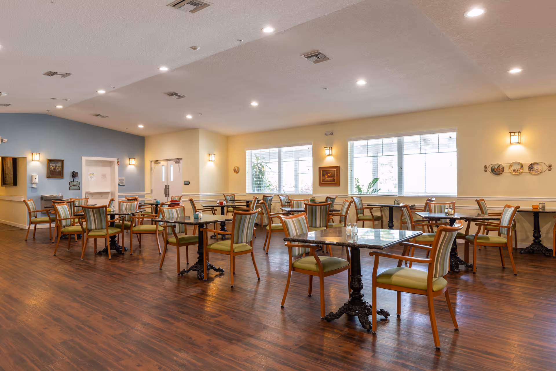 A spacious dining room with multiple tables and chairs arranged neatly. The room has wooden flooring, large windows letting in natural light, and soft yellow and blue walls. Wall-mounted lights and ceiling spotlights illuminate the area, creating a warm and inviting atmosphere.