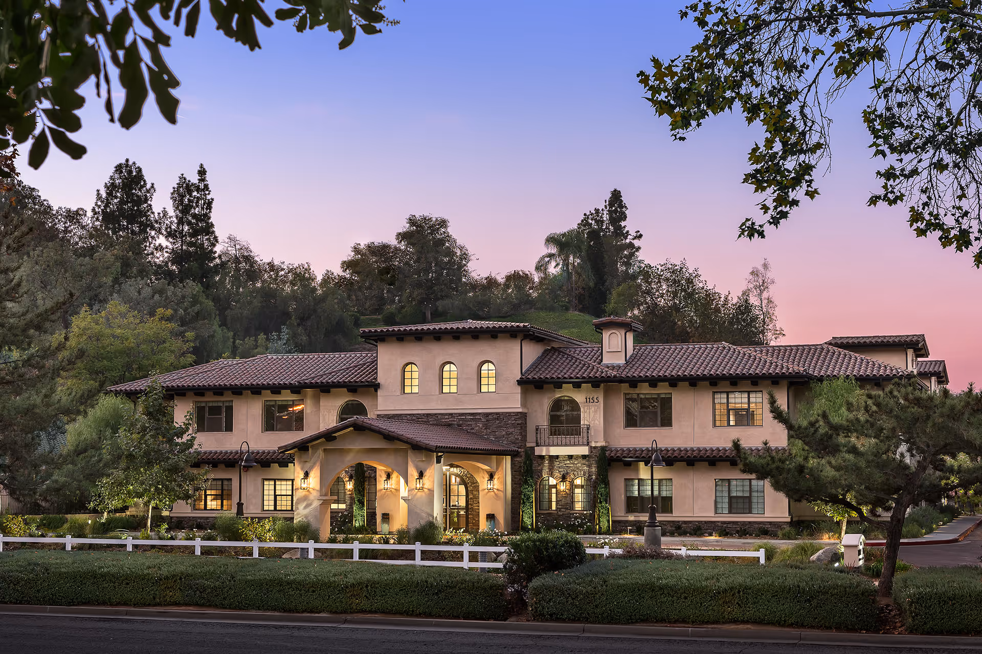 Exterior view of a two-story senior living facility building at dusk with warm lights glowing from the windows. The building has a tiled roof, arched windows, and an entrance with a covered porch. Trees and shrubs surround the building, and a white fence lines the front.