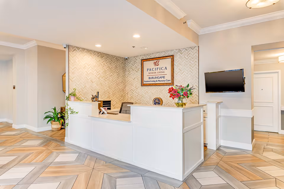 Reception area of Burlingame Senior Living with a white front desk, a wall-mounted TV, decorative plants, and a framed sign that reads 'Pacifica Senior Living Burlingame Assisted Living & Memory Care'. The floor has a patterned wood design and the walls are painted in neutral tones.
