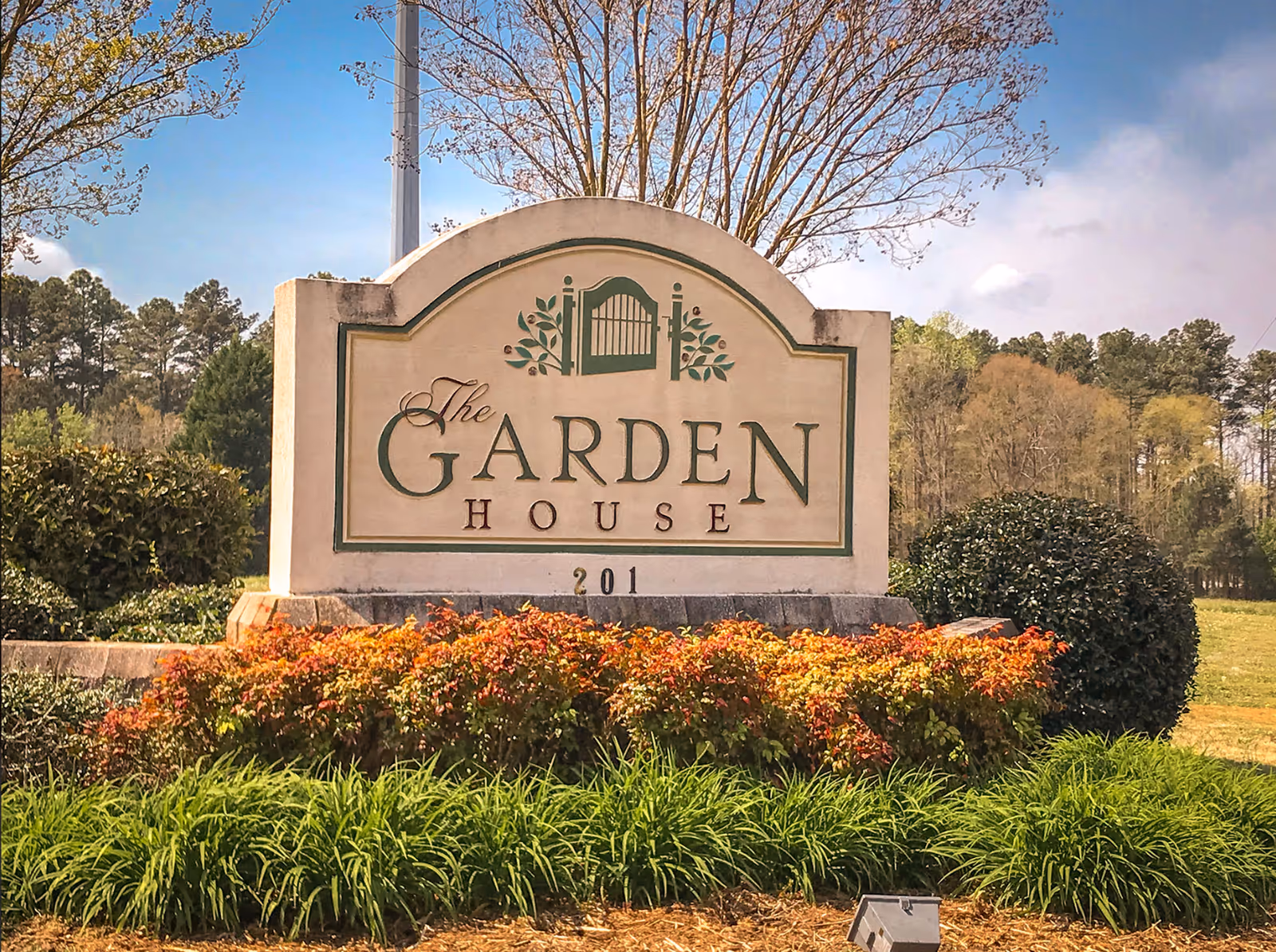 Stone sign for The Garden House with green and brown lettering, surrounded by bushes and plants, with trees and a blue sky in the background.