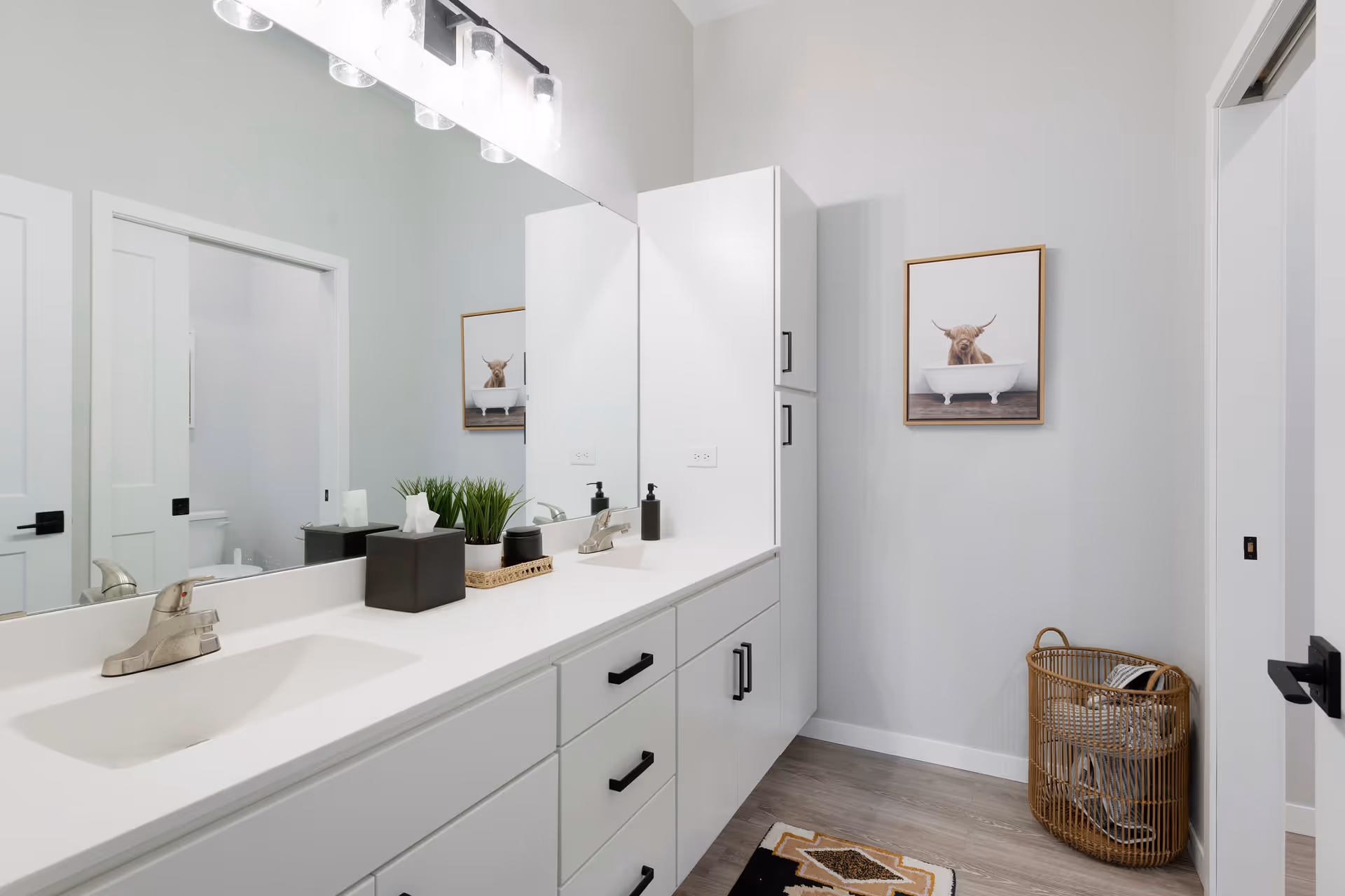 A modern bathroom with a white double sink vanity featuring black handles, a large mirror above the sinks, and a light fixture with four bulbs. There is a decorative plant and soap dispenser on the countertop. On the right wall, there is a framed picture of a cow in a bathtub and a wicker laundry basket with towels inside. The floor is wood-style laminate with a small patterned rug.