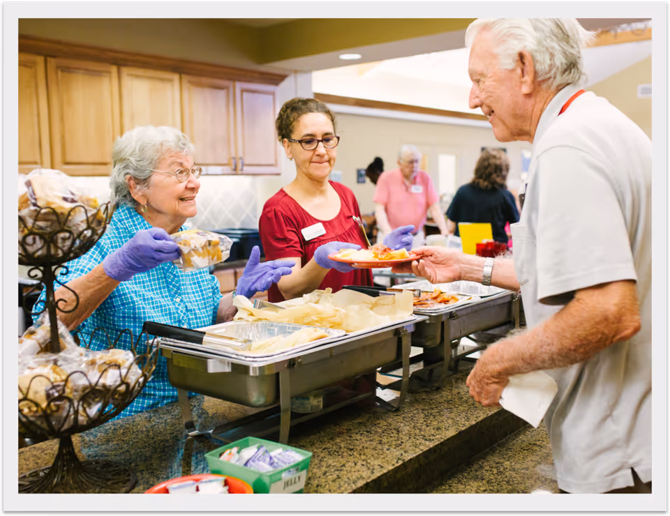 Staff and residents serving and receiving food at a buffet counter in a dining area.