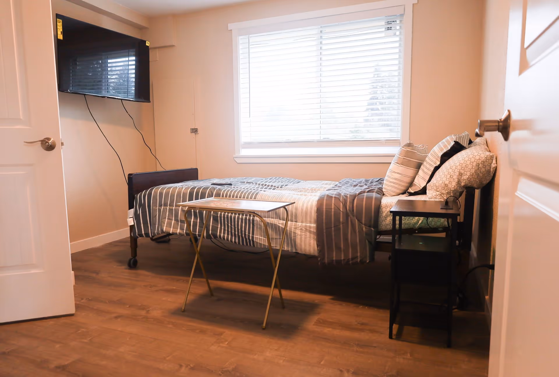 Small furnished bedroom with a single bed, pillows, a bedside table, a wall-mounted TV, and a window with blinds.