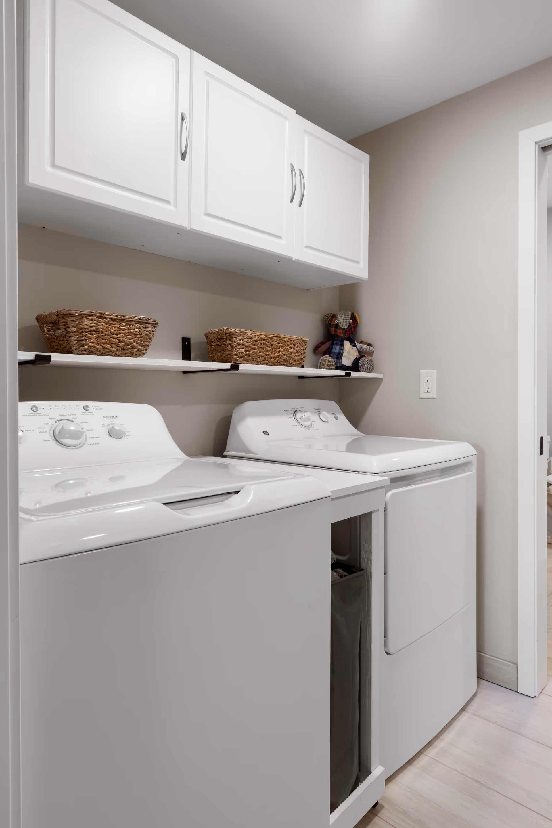 Laundry room with a white washing machine and dryer side by side. Above them is a white cabinet and a shelf holding two woven baskets and a small stuffed bear. The walls are painted light beige and the floor is light-colored wood.